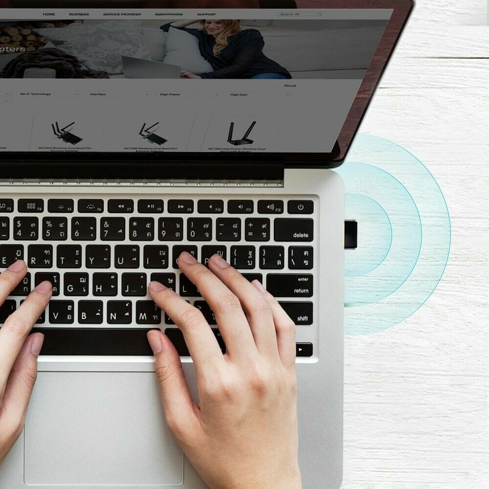 Une personne utilise un ordinateur portable argenté sur un bureau en bois blanc. On voit ses mains sur le clavier et un signal sans fil émis par un petit adaptateur USB branché sur le côté.