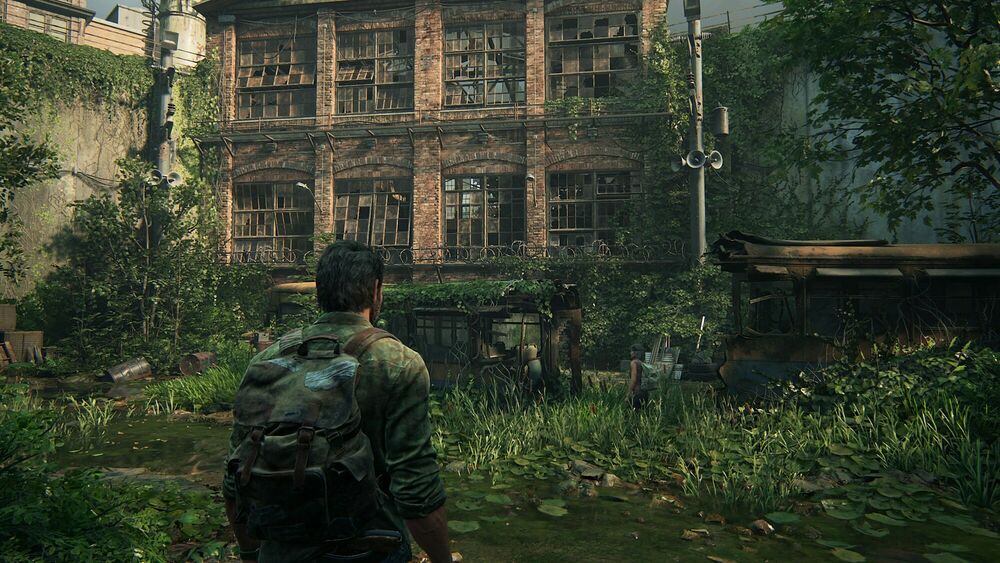 Image de Gameplay de The Last Of Us Part 1. On y voit Joël en vue 3eme personne, seul. Il se situe dans ce qui ressemble à une cour intérieure, et à face à lieu un grand bâtiment ressemblant à une usine, dont la façade et les alentours ont été envahis par la végétation.