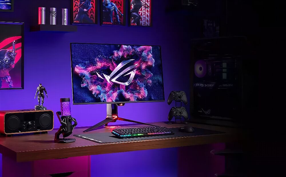 Un setup gaming avec un écran ASUS affichant un fond d'écran coloré, un clavier rétroéclairé, un micro, une enceinte et deux manettes sur un bureau en bois, le tout éclairé par des lumières violettes.