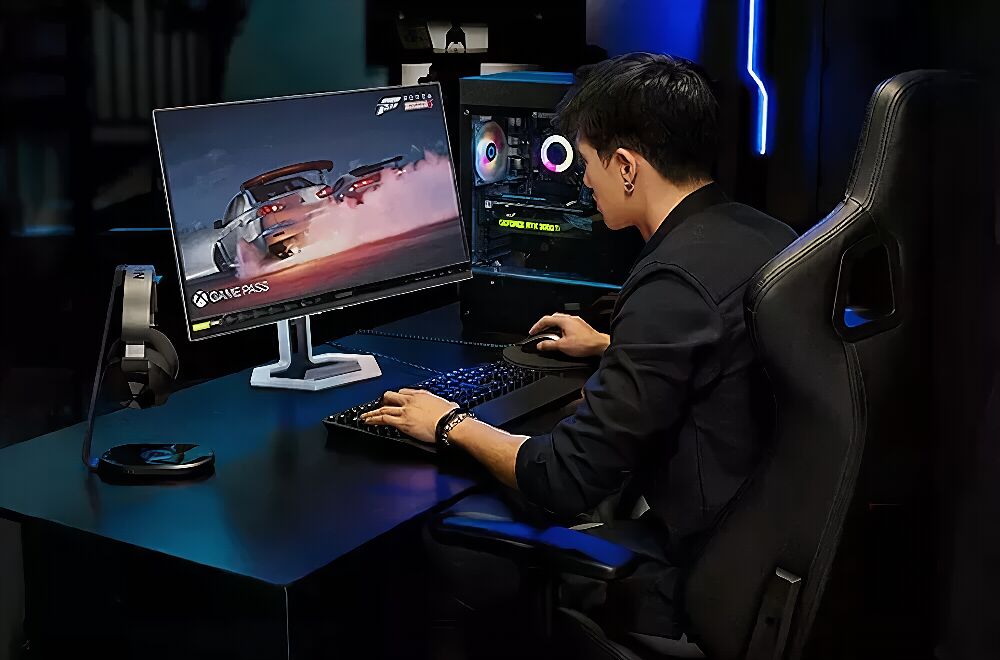 Un homme est assis à un bureau de gaming devant un écran affichant un jeu de course, un clavier, une souris et un casque audio. Le setup comprend également un PC avec un éclairage RGB.