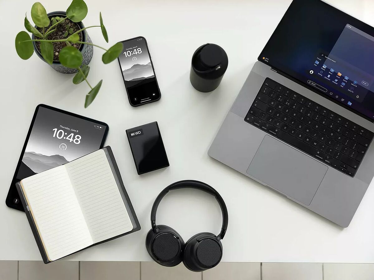 Vue de dessus d'un bureau blanc avec un ordinateur portable, un smartphone, une tablette, un casque audio noir, un carnet ouvert, une plante en pot et un disque dur externe WD.