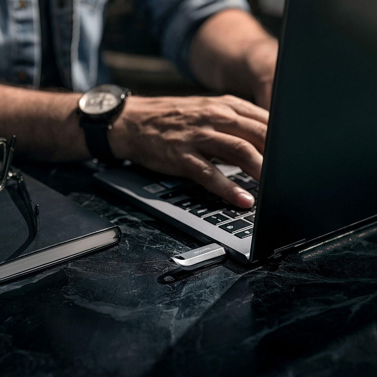 Une personne utilise un ordinateur portable sur une table en marbre, avec une clé USB SanDisk branchée sur le côté de l'appareil.