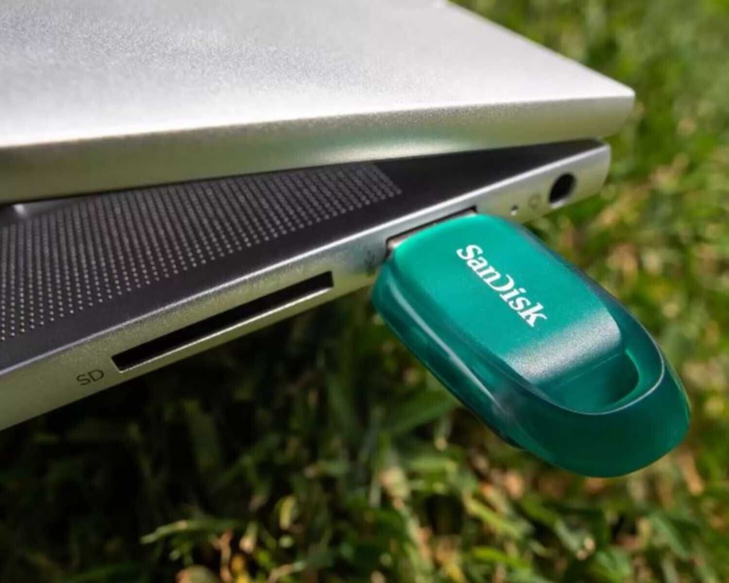 Une clé USB SanDisk de couleur verte est insérée dans le port USB d'un ordinateur portable argenté, posé sur de l'herbe.