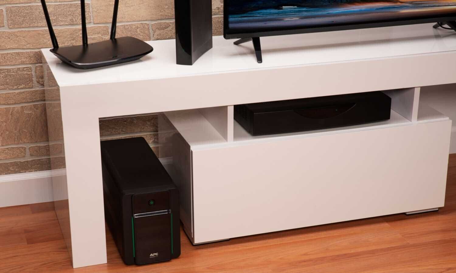 Un onduleur APC noir placé sous un meuble TV blanc à côté d'un boîtier de décodeur.