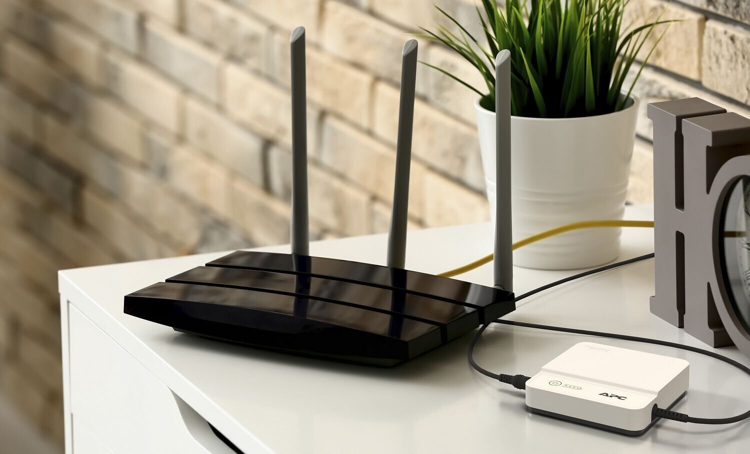 Un routeur Wi-Fi noir avec trois antennes, posé sur un bureau à côté d'une plante en pot et d'un petit boîtier APC.