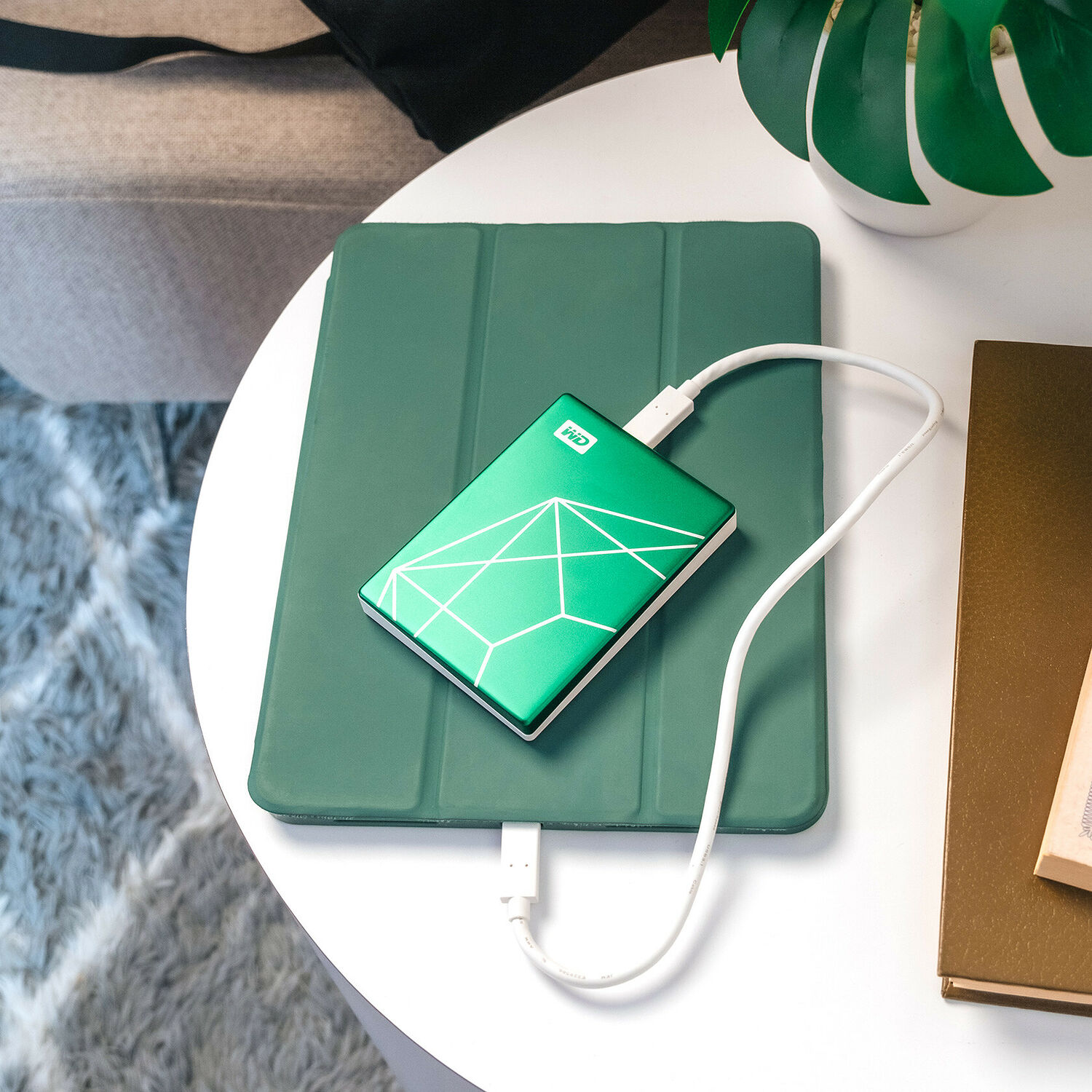Un disque dur externe WD de couleur verte avec un motif géométrique blanc, relié par un câble blanc à une tablette posée sur une table.
