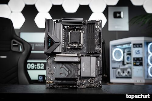 Carte mère Gigabyte Eagle avec dissipateurs thermiques noirs et détails gris, présentée sur un bureau devant un setup gaming.
