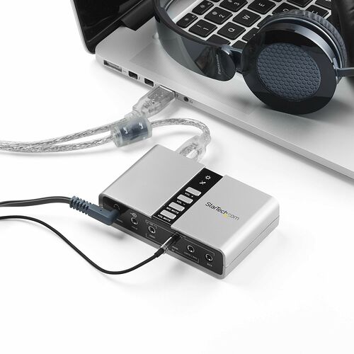 Un adaptateur audio StarTech.com relié à un ordinateur portable, avec un casque branché sur l'appareil. L'adaptateur possède des boutons de contrôle du volume et du micro.