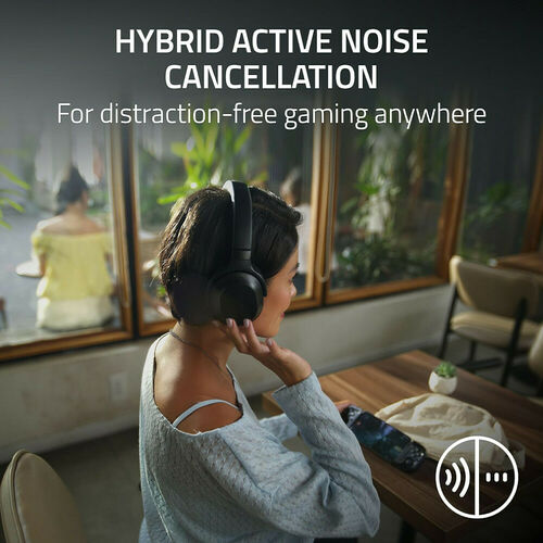 Une femme portant un casque audio noir avec réduction active du bruit hybride, assise à une table en train de regarder son smartphone.