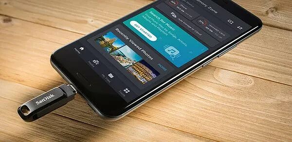 Une clé USB SanDisk branchée sur un smartphone noir posé sur une table en bois. L'écran du téléphone affiche une interface de gestion de fichiers et de musique.