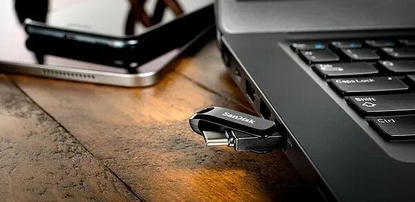 Une clé USB SanDisk branchée sur le port latéral d'un ordinateur portable noir, posé sur une table en bois à côté d'un smartphone.