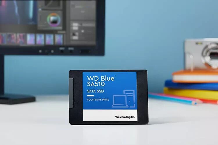 Un SSD SATA Western Digital Blue posé sur un bureau devant un écran et des livres.