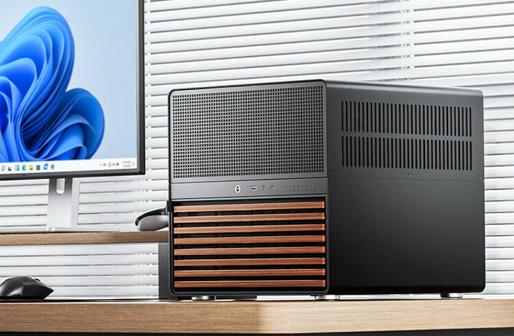 Un PC compact noir avec une façade en lattes de bois, posé sur un bureau à côté d'un écran affichant Windows.