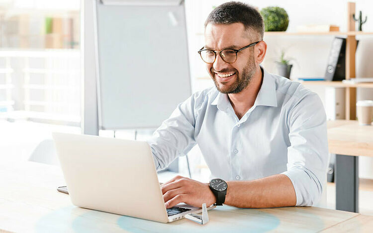 Un homme souriant portant des lunettes et une chemise claire travaille sur un ordinateur portable sur un bureau dans un bureau lumineux.