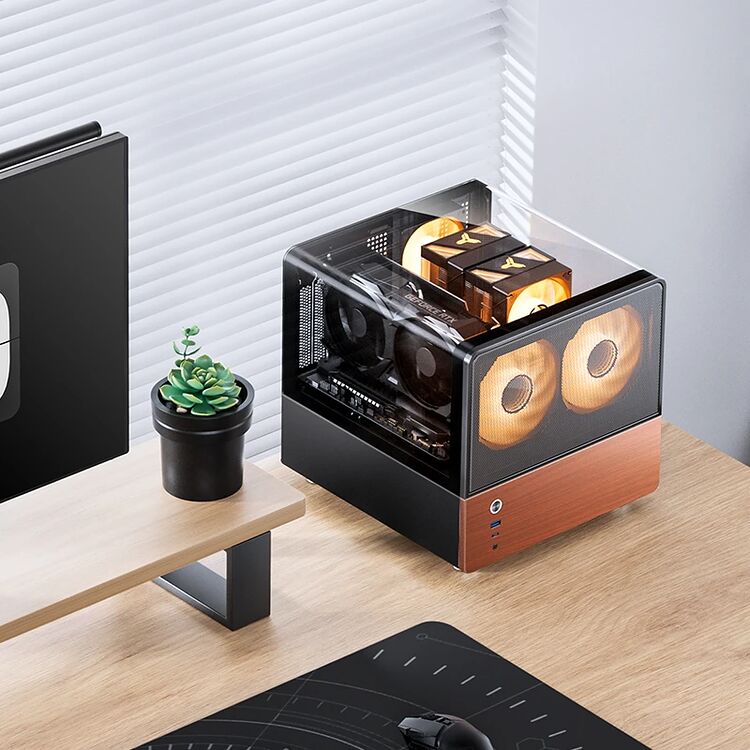 Un petit PC compact avec un boîtier noir et des panneaux latéraux transparents, posé sur un bureau à côté d'un écran et d'une petite plante en pot.