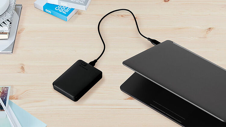 Un disque dur externe noir relié par un câble USB à un ordinateur portable gris sur un bureau en bois clair.