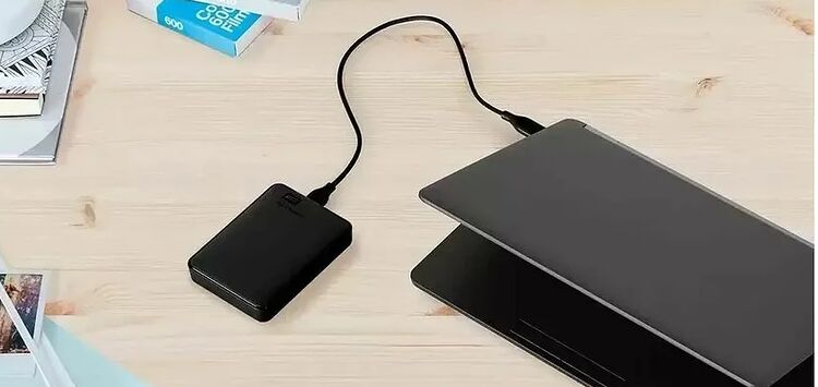 Un disque dur externe noir est relié par un câble USB à un ordinateur portable gris posé sur un bureau en bois clair.