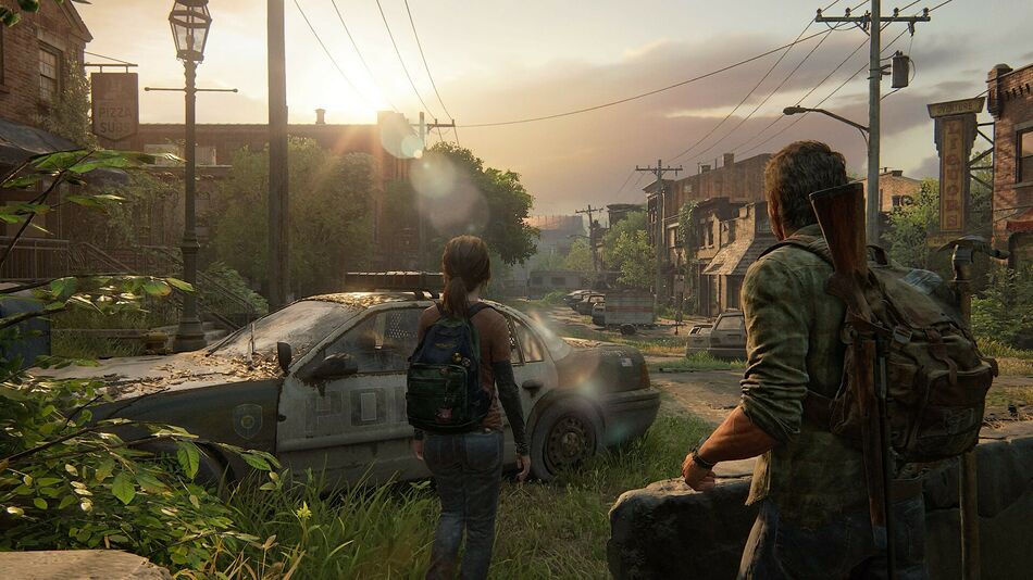 Screenshot tiré du jeu The Last Of Us Part 1. On y voit Joël et Ellie, les deux prtotagonistes du jeux, de dos, regardant au loin le bout d'une rue. La rue à l'air de se trouver dans une zone industrielle où la végétation à repris le dessus.