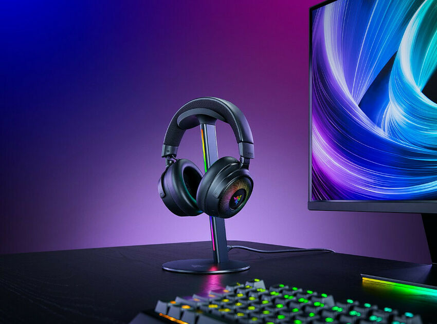 Un casque Razer posé sur son support de rangement, à côté d'un écran d'ordinateur et d'un clavier rétroéclairé sur un bureau.