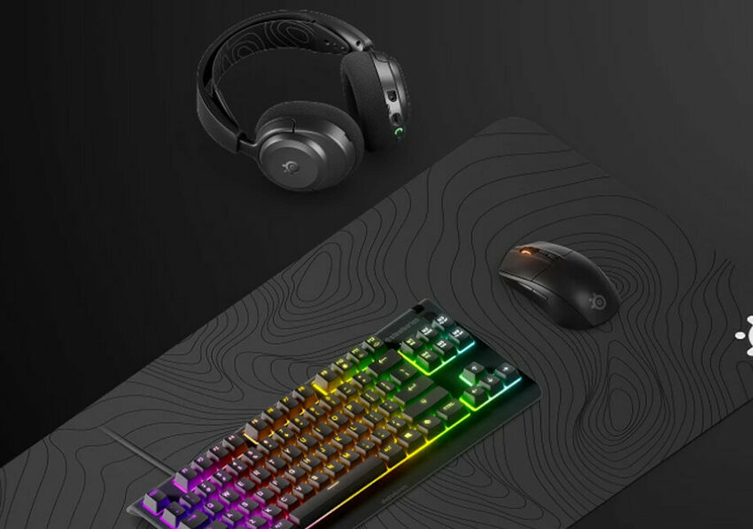 Un ensemble de périphériques gaming comprenant un clavier rétroéclairé, une souris et un casque SteelSeries, le tout disposé sur un tapis de souris noir à motifs de lignes topographiques.
