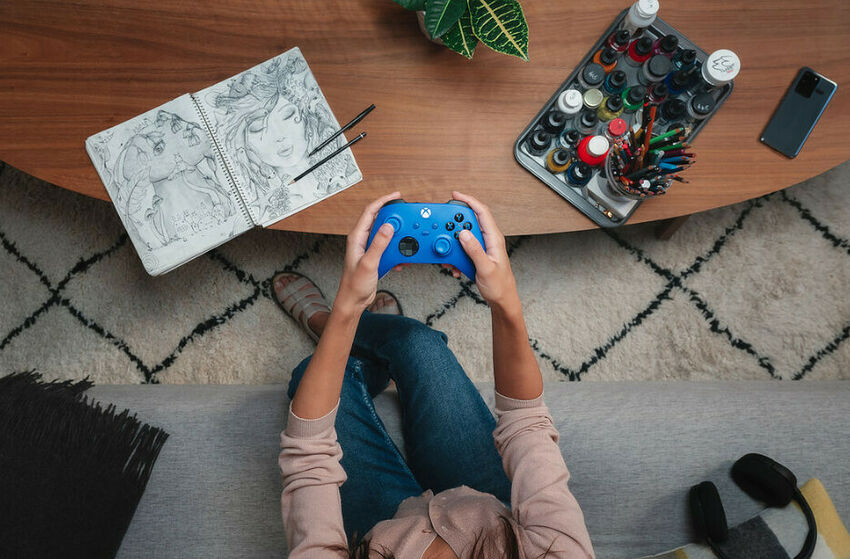 Vue de dessus d'une personne assise sur un canapé jouant avec une manette bleue Xbox, avec un carnet de dessins et des feutres sur une table en bois.