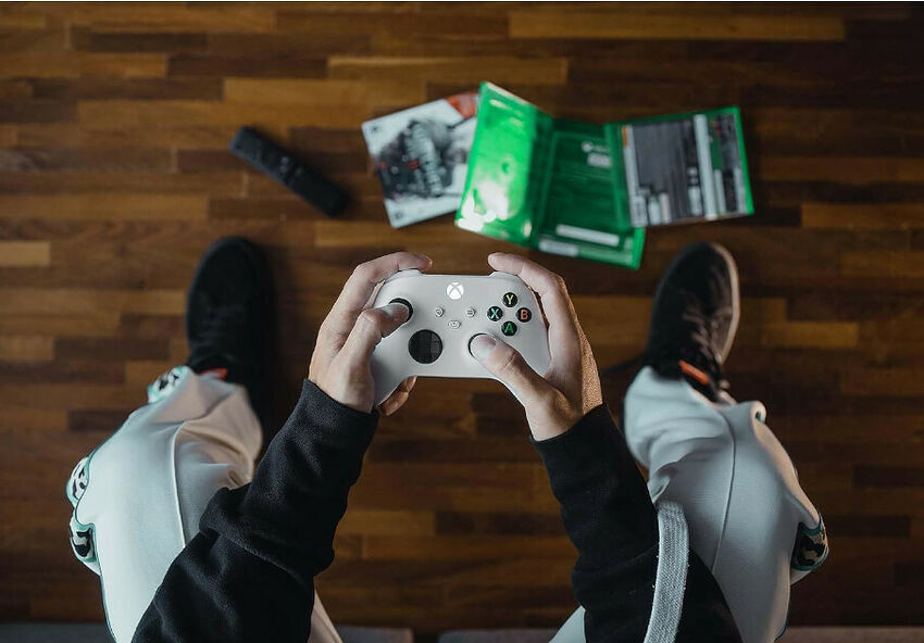 Vue de dessus d'une personne assise par terre tenant une manette blanche Xbox, avec des jeux vidéo et une télécommande posés sur le parquet.