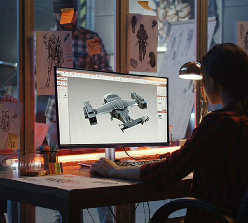Une personne travaille sur un ordinateur de bureau affichant une modélisation 3D d'un vaisseau spatial, dans un bureau décoré de croquis et d'illustrations.