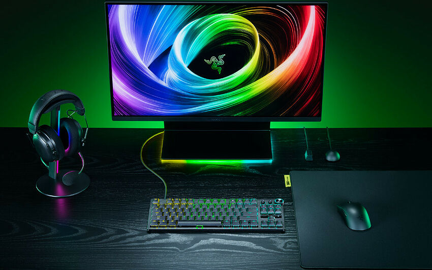 Un setup gaming comprenant un écran Razer avec un fond d'écran coloré, un clavier mécanique rétroéclairé, une souris et un casque audio posé sur son support, le tout sur un bureau noir avec un éclairage RGB vert.