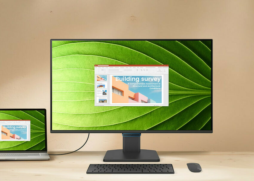 Un écran de bureau affichant une image de feuille verte, accompagné d'un clavier et d'une souris sans fil noirs sur un bureau en bois. Un ordinateur portable est également visible sur la gauche.