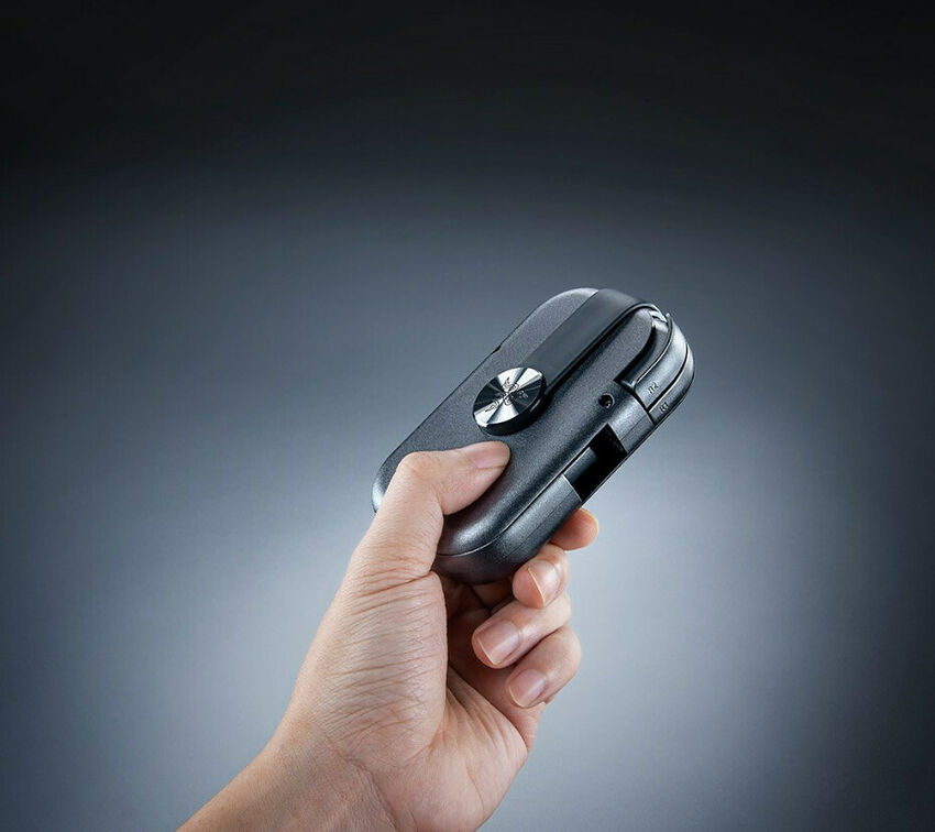 Une main tient un petit appareil électronique noir de forme ovale avec un bouton central argenté.