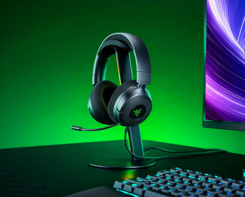 Casque Razer posé sur son support sur un bureau, avec un clavier rétroéclairé et un écran en arrière-plan. L'ensemble est éclairé par une lumière verte.