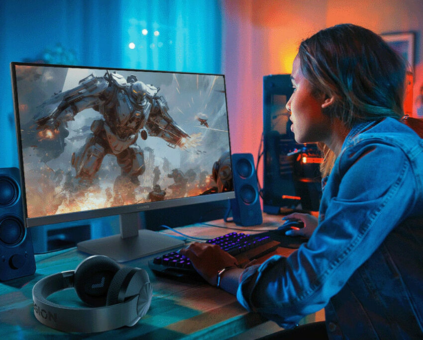 Une femme joue à un jeu vidéo sur un écran Lenovo. Sur son bureau, on voit également un clavier rétroéclairé et un casque audio.