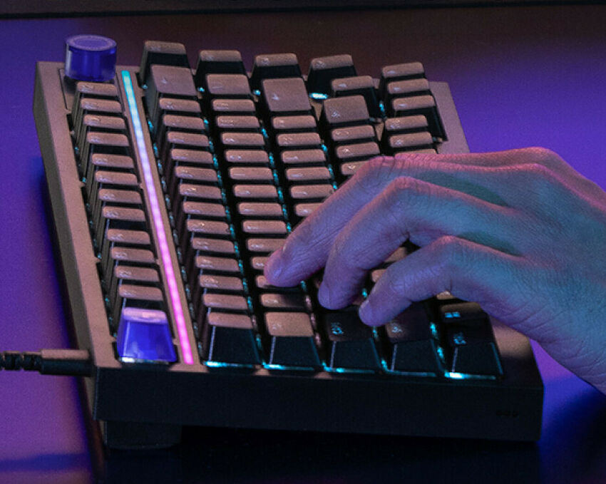 Une main tape sur un clavier mécanique compact avec un rétroéclairage LED coloré. Le clavier présente une barre lumineuse verticale sur le côté gauche.