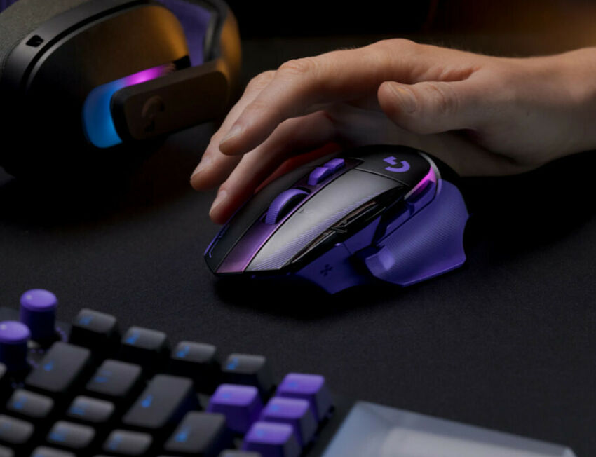 Une main manipule une souris gaming Logitech de couleur violette et noire avec un éclairage RGB, posée sur un tapis de souris à côté d'un clavier et d'un casque.