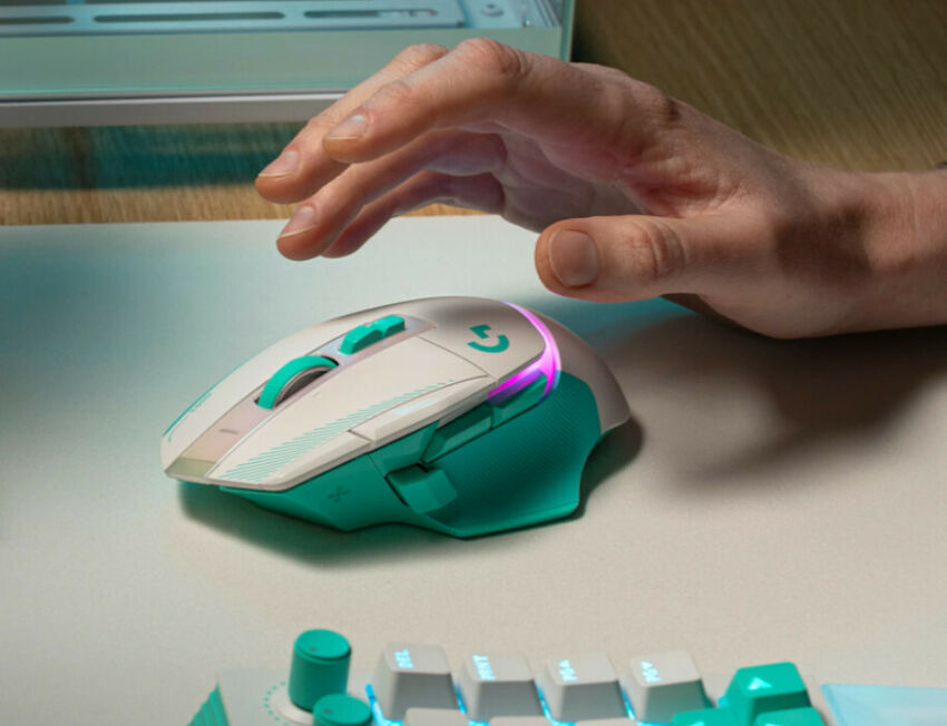 Une main manipule une souris gaming blanche et turquoise avec un éclairage RGB violet. On aperçoit également une partie d'un clavier assorti sur un tapis de souris clair.