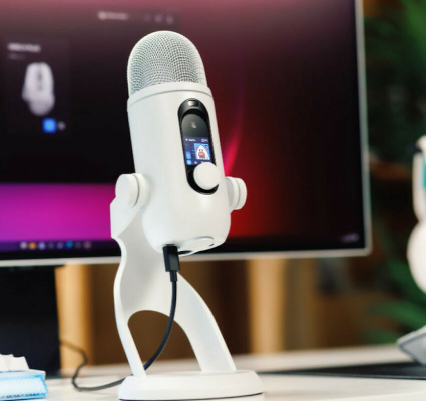 Microphone blanc avec un petit écran LCD et un bouton de contrôle, monté sur un pied de bureau blanc devant un écran d'ordinateur.