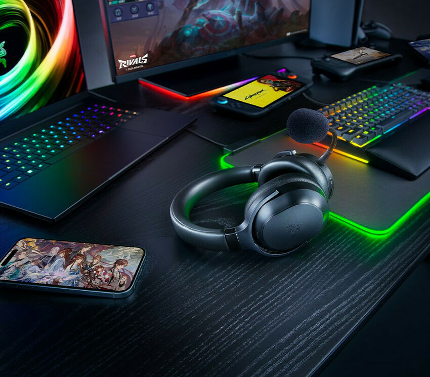 Un casque de gaming noir avec micro, posé sur un bureau avec un clavier rétroéclairé, une souris, un tapis de souris lumineux et deux smartphones.