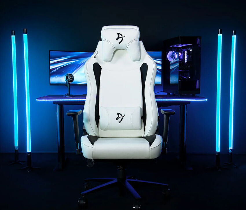 Un siège de gaming blanc et noir de marque DXRacer, placé devant un bureau avec un PC et deux écrans, le tout éclairé par des barres lumineuses bleues.