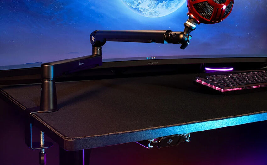 Un bras articulé noir fixé au bord d'un bureau supportant un microphone rouge, devant un écran d'ordinateur et un clavier rétroéclairé.