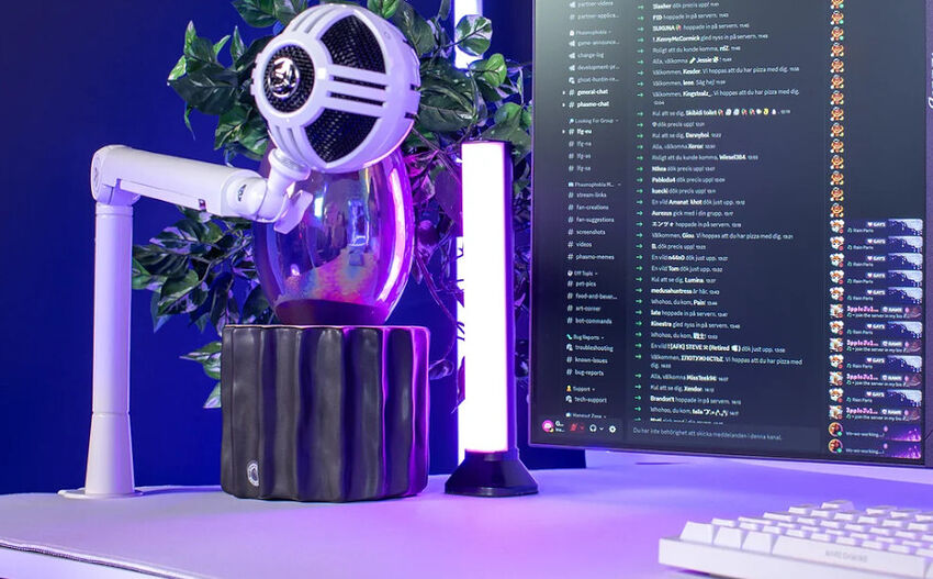 Un microphone blanc monté sur un bras articulé, posé à côté d'une lampe LED verticale sur un bureau, avec un écran affichant une interface de chat en arrière-plan.