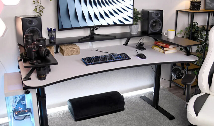 Un bureau de gamer ergonomique avec un plateau blanc incurvé, un clavier mécanique bleu, une souris, un écran, des enceintes noires et un repose-pied noir. Un PC avec éclairage RGB est visible sur le côté.